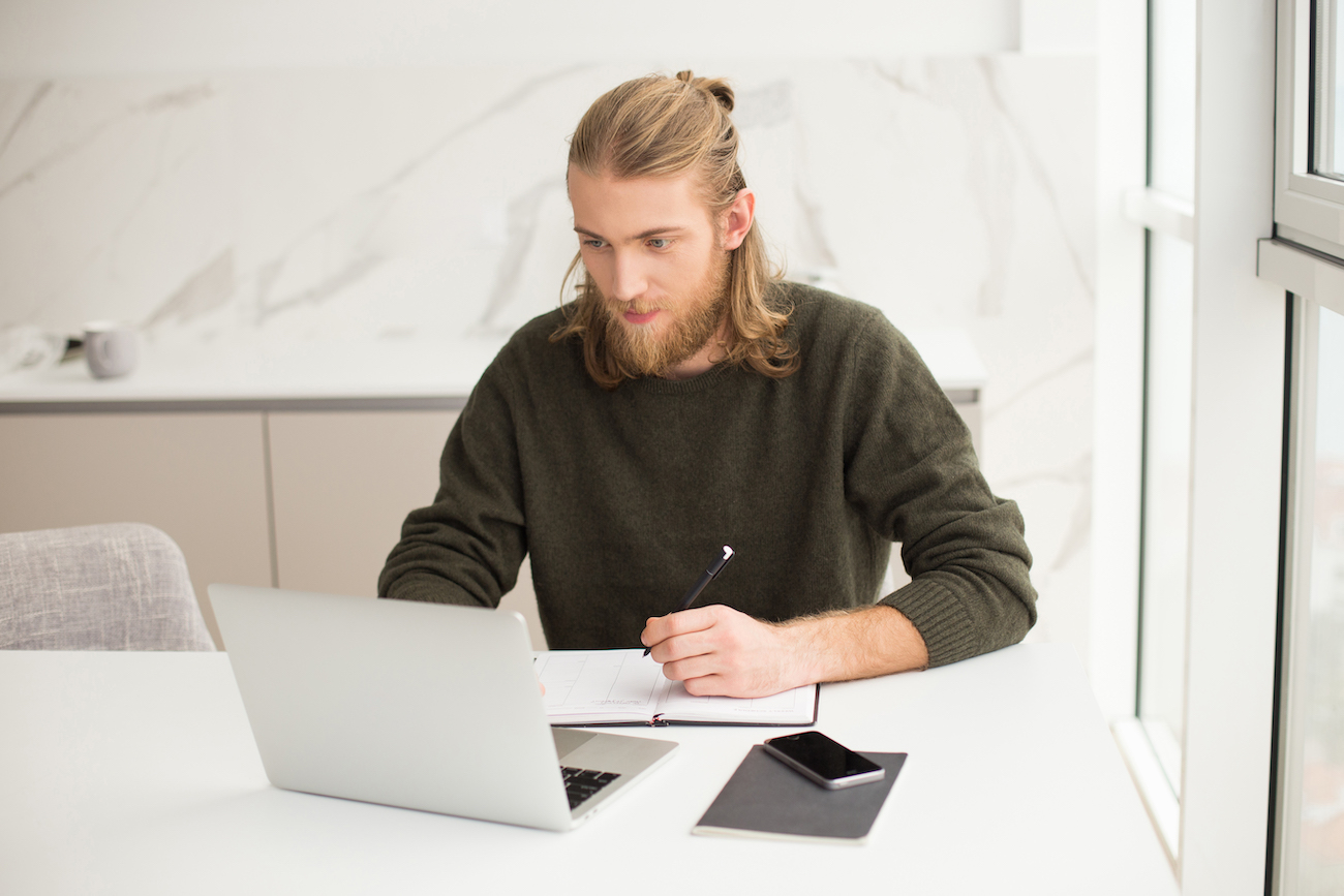 Young man sitting with laptop and making notes in notebook at home