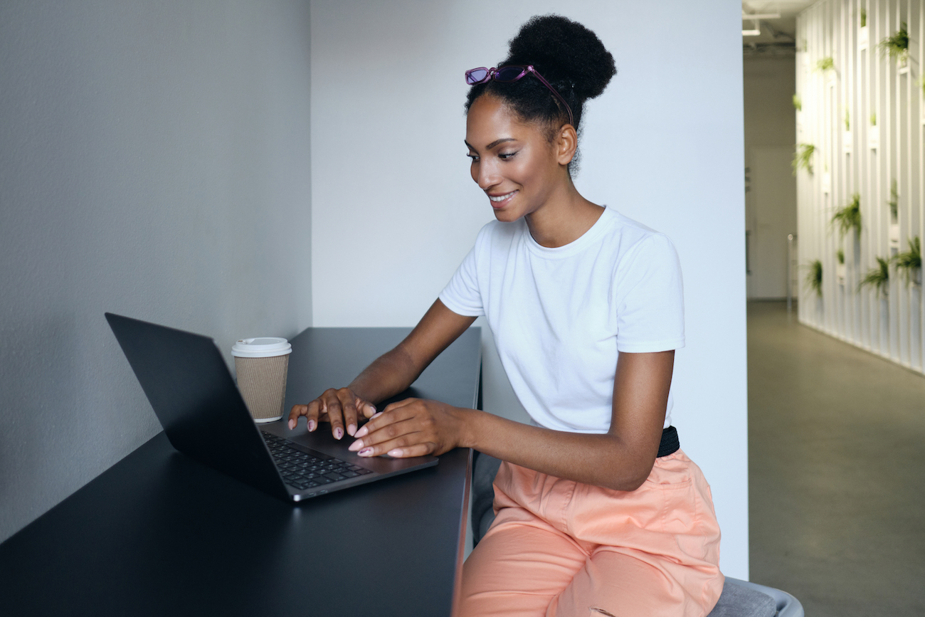 Young pretty smiling casual African American woman working on laptop in co-working space